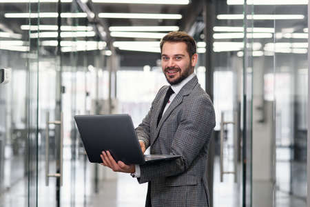 Smiling bearded business man focused on company datas while standing in office corridor and using laptop.の写真素材