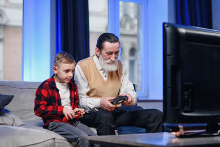 Cute little boy with grandfather sitting on sofa and playing video game with game padの写真素材