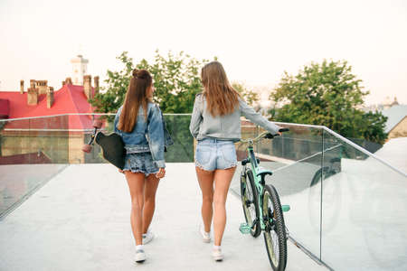 Back view of two adorable best girlfriends in stylish denim jackets and jeans shorts with bicycle and longboard while walking on the street in morning. Friendship concept.の写真素材
