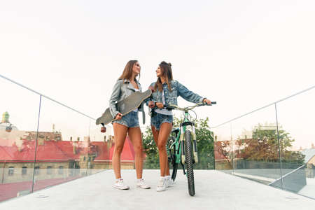 Two young attractive female friends walk with a bike and skateboard on the urban area in sunny...