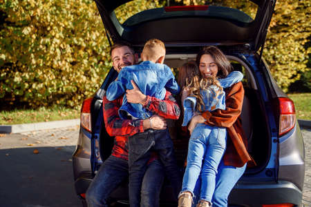 Happy cheerful young parents sitting in the cars trunk and hug their happy kids running to them.の写真素材