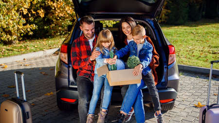 Lovely young parents with their cute kids sitting in the trunk and holding carton box with plants...