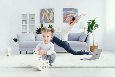 Funny baby girl playing at home while her sporty healthy mommy doing fitness and yoga exercises on the background.の写真素材