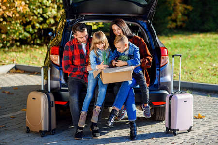 Lovely young parents with their cute kids sitting in the trunk and holding carton box with plants and other home things during moving into new apartment.の写真素材