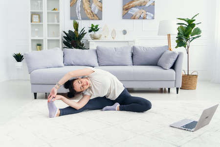 Attractive young professinal female fitness trainer sitting on mat with portable computer having...