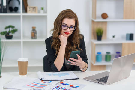 Smiling young business woman in stylish clothes sitting in cozy modern office and browsing something on smartphone.の写真素材