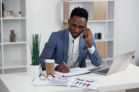 Young black businessman talking on mobile phone and working on laptop in modern white office, copy spaceの写真素材