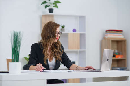 Successful concentrated businesswoman at official clothes sitting in cozy bright office and working with laptop computer. Concept of experienced company manager.の写真素材