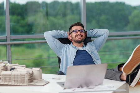 Happy office worker in casual clothes laid his feet on workspace table while dreaming about rest or vacations. Joyful architect relax on the workplace successfully completing his project.の写真素材