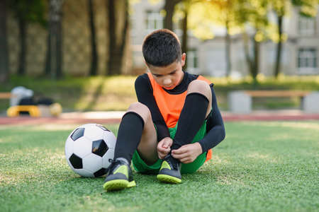 Cute 13-aged football player on artificial green covering of outdoors sport field and tying the shoelace on his boots.の写真素材