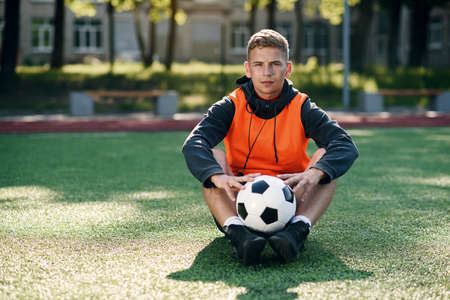 Professional soccer coach in an orange vest holds a ball on hands while sitting at artificial football field.の写真素材