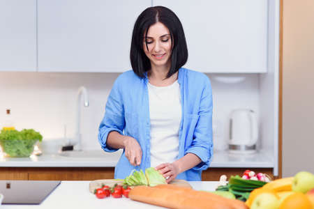 Young woman cuts the cabbage with a knife on a wooden board and preparing salad from vegetables at cozy home kitchen.の写真素材