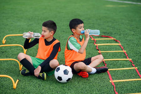 Two teen boys in football uniforms having a rest on sport field with artificial covering and...