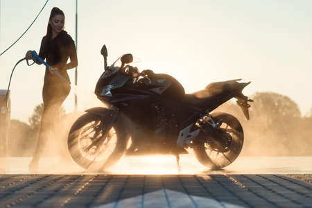 Beautiful pretty girl washes a motorcycle at self-service car wash in the city at sunrise. Taking...