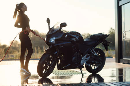 Beautiful stunning girl washes a motorcycle at self-service car wash in the morning. Taking care...