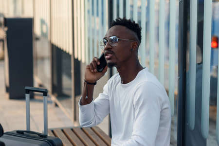 Black male traveler with a suitcase calls a taxi on mobile phone. Attractive tourist speaking on...