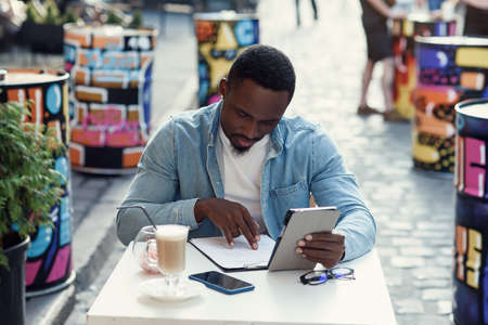 African american business man analyzes paper documents and works on laptop sitting on outdoor cafe...