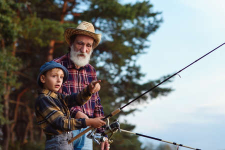 Stylish experienced bearded grandpa in straw hat fishing together with his curious grandchild on the lake at sunset.の写真素材