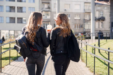 Back view of teen pretty girlfriends with backpacks walking on the street.