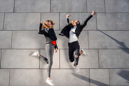 Two beautiful teenage girlfriends jump and having fun near grey concrete wall. Leisure time with best friends.の写真素材