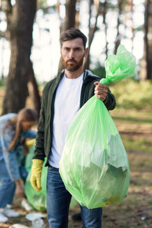 Good-looking bearded man shows a full rubbish pack in background of his friends volunteers picking up rubbish at park.の写真素材