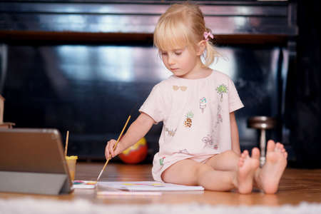 Little funny girl painting with paintbrush and colorful paints sitting on the floor at home. Kids...