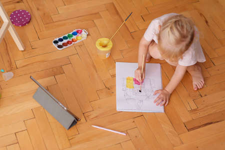 Little funny girl painting with paintbrush and colorful paints sitting on the floor at home. Top view.の写真素材