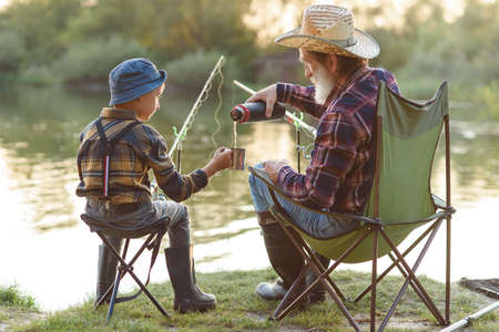 Careful grandpa with grey beard in straw hat sitting on folding chairs with his grandchild on fishing and drinking tea.の写真素材