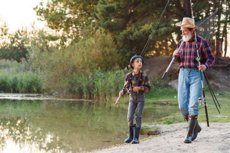 Happy excited boy with his experienced old grey-bearded granddad going near the lake with rods while fishing at sunset.の写真素材