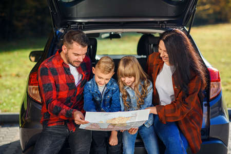 Satisfied pleasant family of handsome father, pretty mother and lovely kids which are looking on the road map while sitting in the cars trunk at joint family vacation.の写真素材