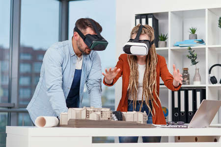 Group of male and female engineers at VR goggles working with mock-up of a house in modern office.
