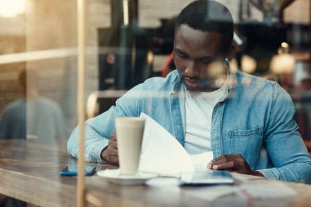 Concentrated black male entrepreneur reads paper documents sitting in cafe with coffee near window.の写真素材