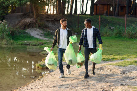 Handsome mixed race active friends carrying plastic bags near the lake after cleanup surrounding territory from rubbish.の写真素材
