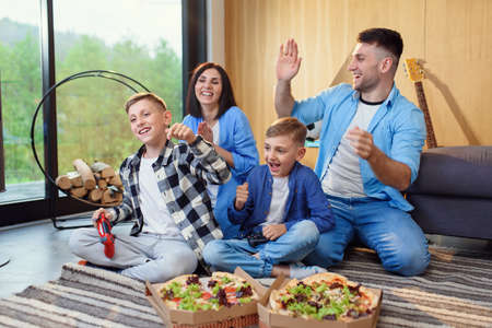 Happy stylish family sitting on floor, playing video games with gamepads and eating tasty pizza.の写真素材