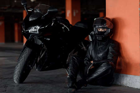 Stylish young woman motorcycle rider with beautiful eyes in black protective gear and full-face helmet near her bike on underground parking.の写真素材