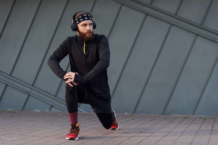 Handsome young man with headphones doing stretching and warm-up exercises before running while...