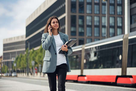 Beautiful formal dressed business woman in suit with a laptop and notebook outdoor. Brunette caucasian female, standing near office.の写真素材