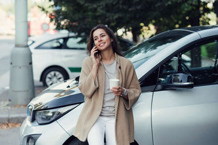 Young beautiful smiling woman with cup of coffee speaks on smartphone near her electric vehicle.の写真素材
