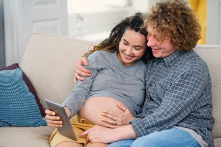 Good-looking pregnant couple relaxing on the comfortable couch at home and watching tv shows on tablet pc.の写真素材