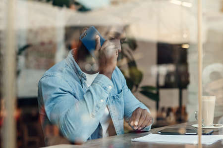 Handsome stylish bearded dark-skinned man sitting near street cafe window and talks on smartphone with business partner.の写真素材
