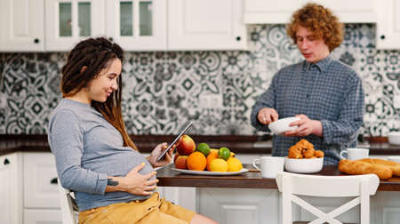 Joyful pregnant woman with dreadlocks drinks tea and watching her curly husband cooking breakfast for her in kitchenの写真素材
