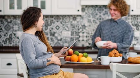 Joyful pregnant woman with dreadlocks drinks tea and watching her curly husband cooking breakfast for her in kitchenの写真素材