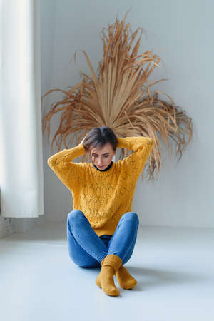 Thoughtful casual young woman in jeans and yellow wool sweater sitting cross-legged on the floor in white interior.の写真素材