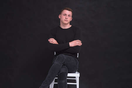 Portrait of a cute young guy sitting on a chair on a black background in different poses. He sits...
