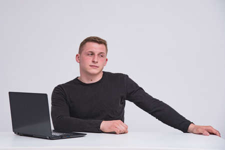 Portrait of a cute young guy with a laptop on a white background sitting at a table. He sits right in front of the camera smiling and looks happyの写真素材