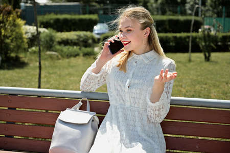 Model posing with phones and smiling in sunny weather outdoors. A photo of a pretty blonde in a park...