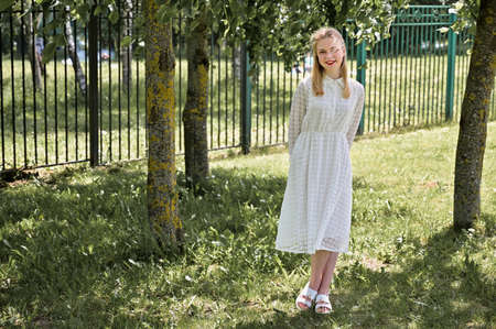 Full length portrait of a nice blonde girl smiling in a park. A Caucasian model in the background of trees stands in sunny weatherの写真素材