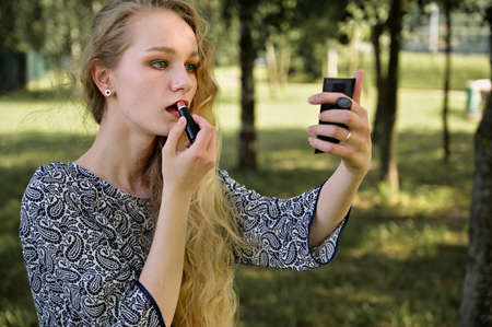 Portrait of a caucasian blonde girl doing makeup in the park. Photo of a pretty young woman with...