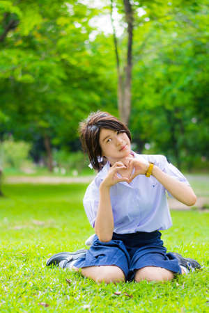 Cute Thai schoolgirl is sitting on the grass and doing heart symbol, spreading love to everyone の写真素材