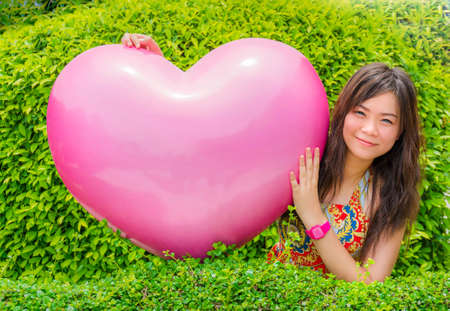 A cute Asian  Thai  girl is hugging giant pink heart in green leaves backgroundの写真素材